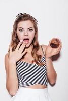 mulher jovem e bonita presa segurando um cupcake e cobrindo a boca com a mão em pé contra um fundo branco foto
