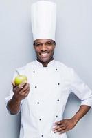 um ingrediente importante para salada de frutas. confiante jovem chef africano em uniforme branco esticando a maçã verde e olhando para a câmera com sorriso em pé contra um fundo cinza foto