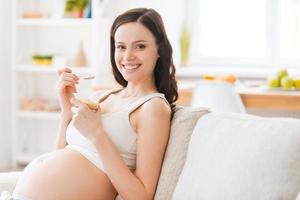 comida saudável para meu bebê jovem grávida alegre comendo iogurte e sorrindo para a câmera enquanto está sentado no sofá foto