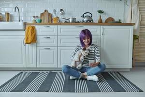 jovem carregando cachorrinho e usando telefone inteligente enquanto está sentado no chão na cozinha foto