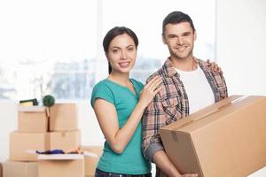 mudança para uma nova casa. alegre casal jovem perto um do outro e sorrindo para a câmera enquanto homem segurando uma caixa de papelão foto