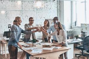 brindando seu sucesso. grupo de jovens empresários brindando uns aos outros e sorrindo em pé atrás da parede de vidro na sala de reuniões foto