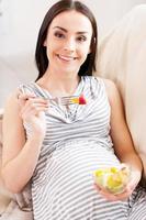 Alimentação saudável. vista superior da linda mulher grávida comendo uma salada de frutas e sorrindo para a câmera enquanto estava deitado no sofá foto