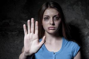 pare de ferir mulher jovem mulher espancada olhando para a câmera e esticando a mão em pé contra a parede escura foto