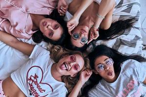 belezas sedutoras. vista superior de quatro mulheres jovens brincalhonas em óculos sorrindo e fazendo uma careta enquanto estava deitado na cama em casa foto