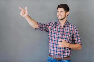 apontando seu anúncio. jovem feliz apontando para longe e sorrindo em pé contra um fundo cinza foto