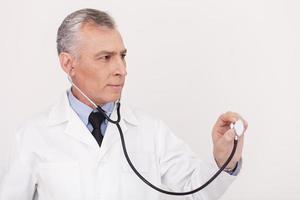 examinando um espaço de cópia. médico sênior de cabelos grisalhos de uniforme olhando para longe e segurando seu estetoscópio estendido em pé isolado no branco foto