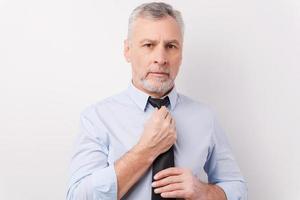 acostumada ao sucesso. homem sênior de cabelos grisalhos confiante em trajes formais, ajustando sua gravata e olhando para a câmera em pé contra um fundo branco foto