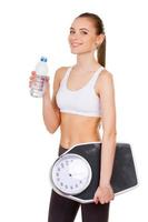 vivendo uma vida saudável. mulher jovem e atraente em roupas esportivas segurando escala de peso e garrafa com água em pé isolado no branco foto
