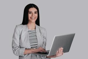 como posso ajudá-la jovem linda em roupas casuais inteligentes, segurando o laptop e olhando para a câmera em pé contra um fundo cinza foto