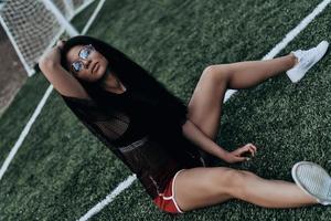 hora de relaxar. mulher jovem e bonita em roupas esportivas, mantendo a mão no cabelo enquanto está sentado no campo de futebol foto