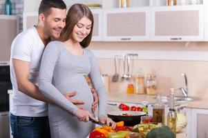 cozinhar alimentos saudáveis. casal jovem feliz cozinhando juntos na cozinha enquanto homem tocando abdômen de sua esposa grávida foto