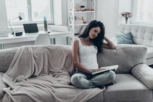 perdido em seu romance. bela jovem lendo um livro enquanto está sentado no sofá em casa foto