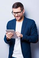 examinando seu novo tablet. jovem alegre trabalhando em tablet digital e sorrindo em pé contra um fundo cinza foto