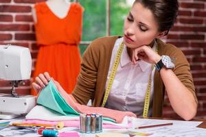 examinando têxteis antes de costurar. designer de moda feminino pensativo examinando têxteis enquanto está sentado em seu local de trabalho e com manequim no fundo foto