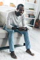 bom fim de semana em casa. jovem africano bonito jogando videogame e sorrindo enquanto está sentado no sofá em casa foto