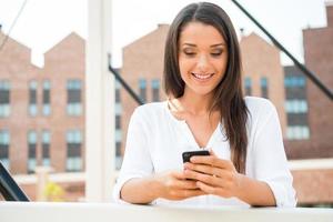 lendo a mensagem dele. bela jovem sorridente segurando o celular e olhando para ele em pé ao ar livre foto