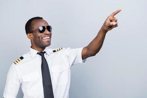 apaixonada pelo céu. alegre piloto africano de uniforme apontando para longe e sorrindo em pé contra um fundo cinza foto