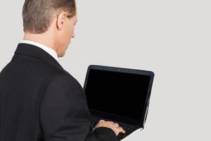 empresário com laptop. vista traseira do homem sênior em trajes formais, digitando algo no laptop em pé contra um fundo cinza foto