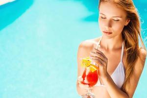 aproveitando o verão. bela jovem de biquíni branco segurando coquetel e mantendo os olhos fechados enquanto relaxa à beira da piscina foto