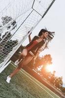 beleza esportiva em movimento. comprimento total de mulher jovem e atraente em biquíni vermelho posando perto da baliza no campo de futebol foto