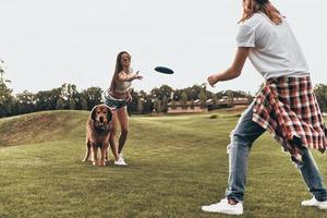 cão lhes traz alegria. comprimento total do belo casal jovem jogando disco de plástico enquanto brincava com seu cachorro ao ar livre foto