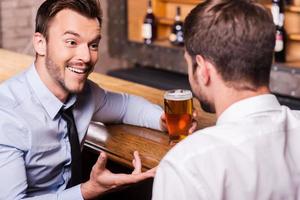 compartilhando cerveja com um bom amigo. dois jovens alegres de camisa e gravata conversando entre si e gesticulando enquanto bebem cerveja no balcão do bar foto