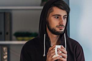 sentindo frustrado. retrato de jovem pensativo na camisa com capuz segurando a xícara de café e desviar o olhar em pé em casa foto