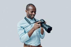 feliz jovem africano usando câmera digital e sorrindo em pé contra um fundo cinza foto