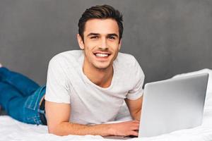 net de surf na cama. jovem alegre olhando para a câmera com sorriso e usando seu laptop enquanto estava deitado na frente na cama contra um fundo cinza foto