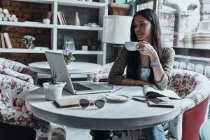 café fresco em seu lugar favorito. mulher jovem e atraente sorridente segurando uma xícara de café e olhando para o laptop enquanto está sentado no restaurante perto da janela foto