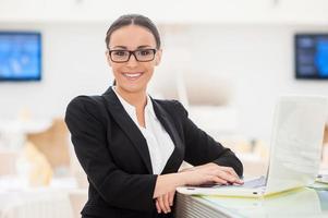 senhora de negócios de sucesso. mulher jovem e bonita em trajes formais trabalhando no laptop e sorrindo enquanto se inclina no balcão do bar foto