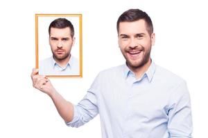 agora estou feliz jovem bonito de camisa segurando uma foto de si mesmo com cara chateada e sorrindo em pé contra um fundo branco