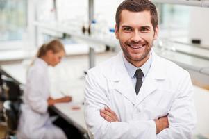 cientista confiante. feliz jovem cientista masculino mantendo os braços cruzados e olhando para a câmera enquanto sua colega trabalhando em segundo plano foto