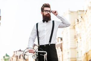 confiante em seu estilo perfeito. vista de ângulo baixo de jovem bonito, inclinando-se para a bicicleta e ajustando os óculos em pé ao ar livre foto