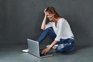 você pode encontrar qualquer coisa online. mulher jovem e atraente em roupas casuais usando computador e sorrindo enquanto está sentado no chão contra um fundo cinza foto