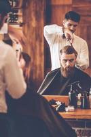 deixando o corte de cabelo perfeito. jovem barbudo cortando o cabelo pelo cabeleireiro enquanto está sentado na cadeira na barbearia na frente do espelho foto