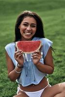 mulher jovem e atraente segurando uma fatia de melancia e sorrindo enquanto está sentado ao ar livre foto