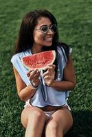 refresco de verão. mulher jovem e atraente em óculos segurando uma fatia de melancia e sorrindo enquanto está sentado na grama foto