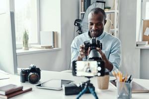 bonito jovem africano de camisa mostrando câmera digital ao fazer vídeo de mídia social foto