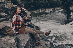 hot tee lida com qualquer problema. lindo casal jovem coberto com cobertor olhando para longe e sorrindo enquanto está sentado nas rochas perto do rio foto