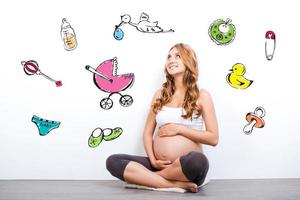 esperando um bebê. mulher grávida sorridente tocando sua barriga e olhando para cima enquanto está sentado contra um fundo branco foto
