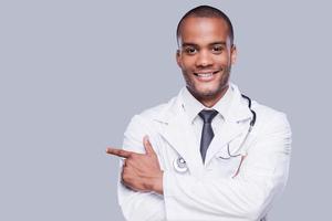 médico masculino confiante. alegre médico africano apontando para longe e sorrindo em pé contra um fundo cinza foto