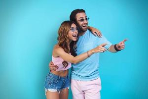 casal surpreso abraçando e apontando para longe em pé contra um fundo azul foto