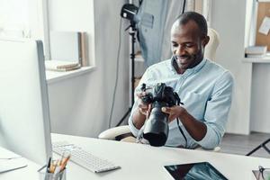 bonito jovem africano segurando a câmera digital e sorrindo enquanto trabalhava no escritório moderno foto