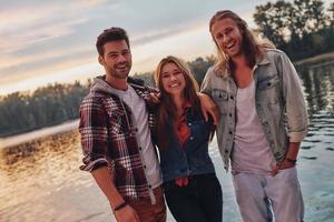 sentindo-se aconchegados juntos. grupo de jovens em roupas casuais sorrindo e olhando para a câmera em pé perto do lago foto