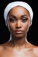 sua perfeição em sua tez. retrato de uma linda mulher africana usando um lenço na cabeça e olhando para a câmera em pé contra um fundo preto foto