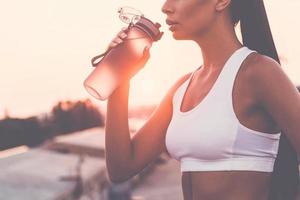 se refrescando. close-up de mulher jovem e bonita em roupas esportivas bebendo água e parecendo cansado em pé na ponte com a luz do sol da noite e vista urbana ao fundo foto