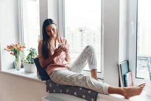 mulher jovem e bonita de pijama aconchegante usando telefone inteligente e sorrindo enquanto descansava no peitoril da janela em casa foto