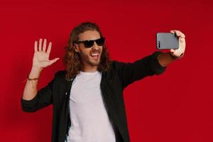 jovem alegre em roupas casuais tomando selfie e sorrindo em pé contra um fundo vermelho foto